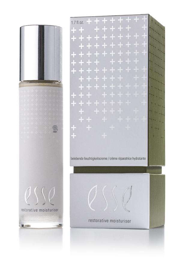 En flaske Restorative Moisturiser med sølvhette står ved siden av den matchende sølvfargede og grønne esken, begge med et minimalistisk design med plusstegn og hvit tekst.