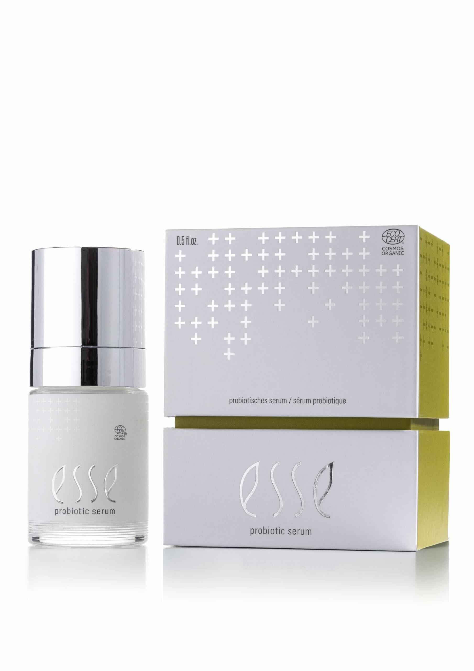 En hvit flaske med Esse Probiotic Serum med sølvglinsende hette vises ved siden av den hvite og gule emballasjen, som har sølvfarget kryssmønster og produktmerkevare.