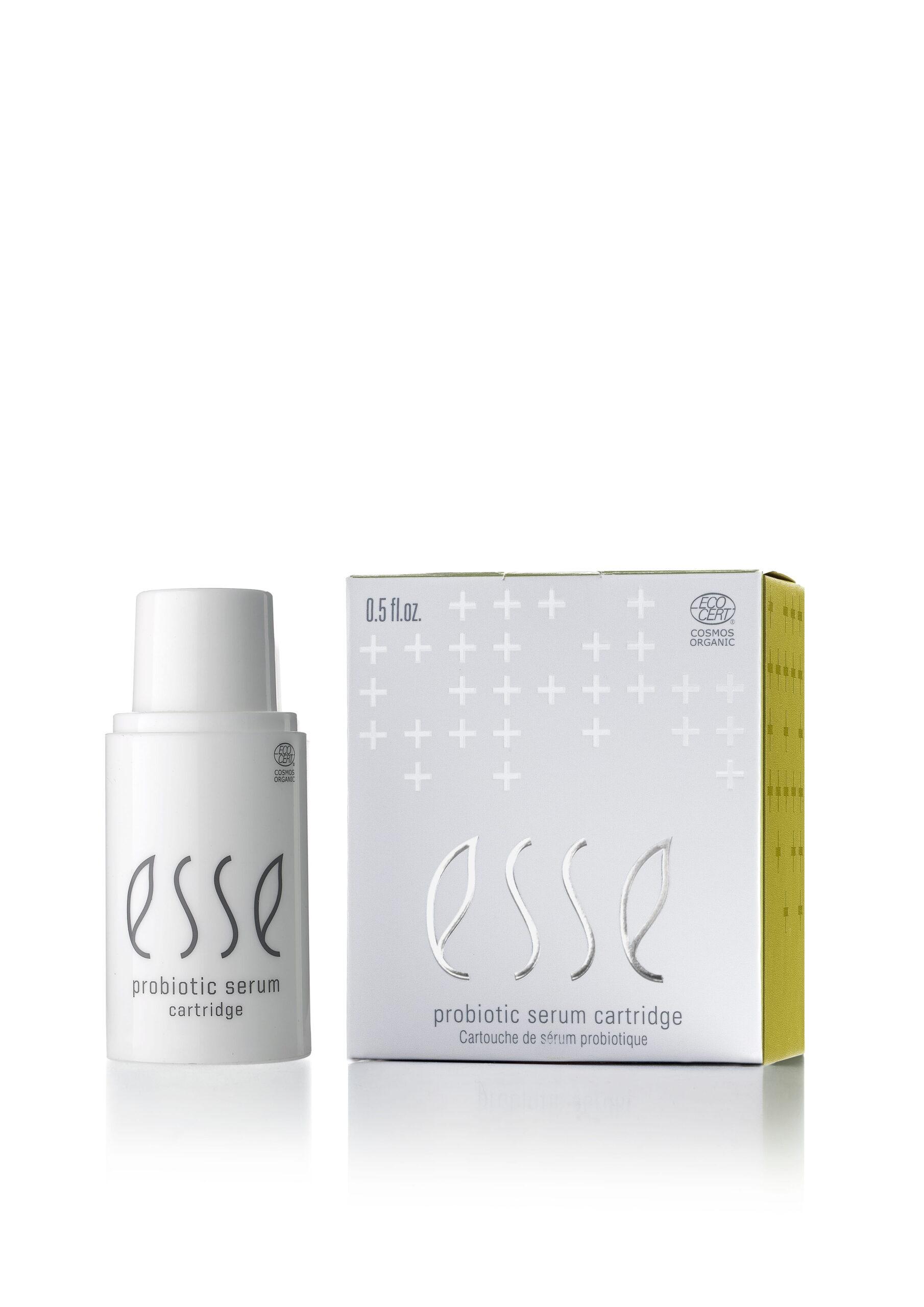 En hvit flaske merket "Esse Probiotic Serum" står ved siden av den matchende esken, som har grå plusstegn, en gul side og "0,5 fl. oz." på toppen.