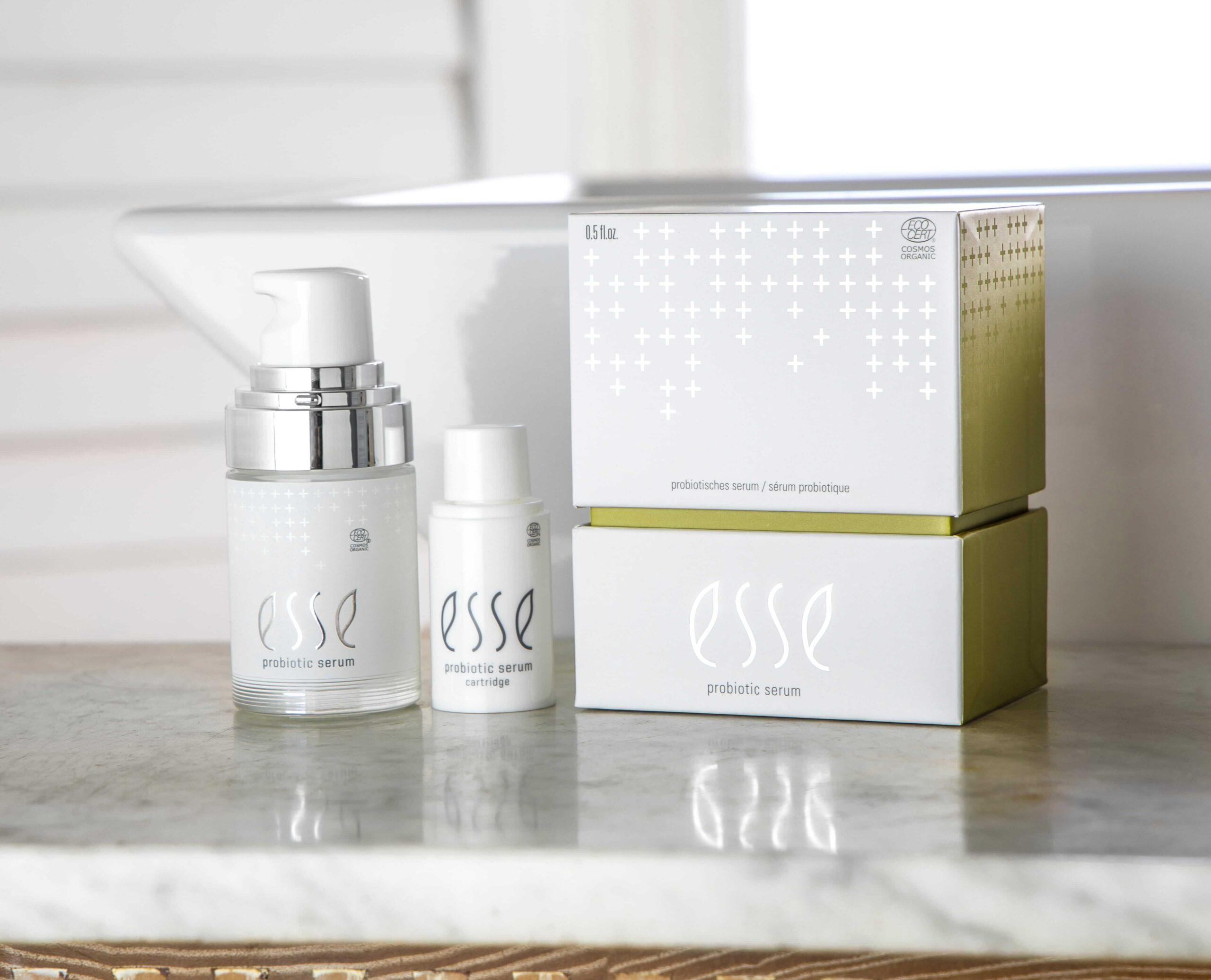 To flasker med Esse Probiotic Serum og produktesken er avbildet på en marmoroverflate med hvit bakgrunn og et vindu i utsikten. Emballasjen har hvite, sølvfargede og lysegrønne aksenter.